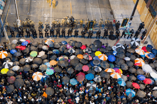 Protesters stand in the rain with umbrellas facing a line of armed riot police