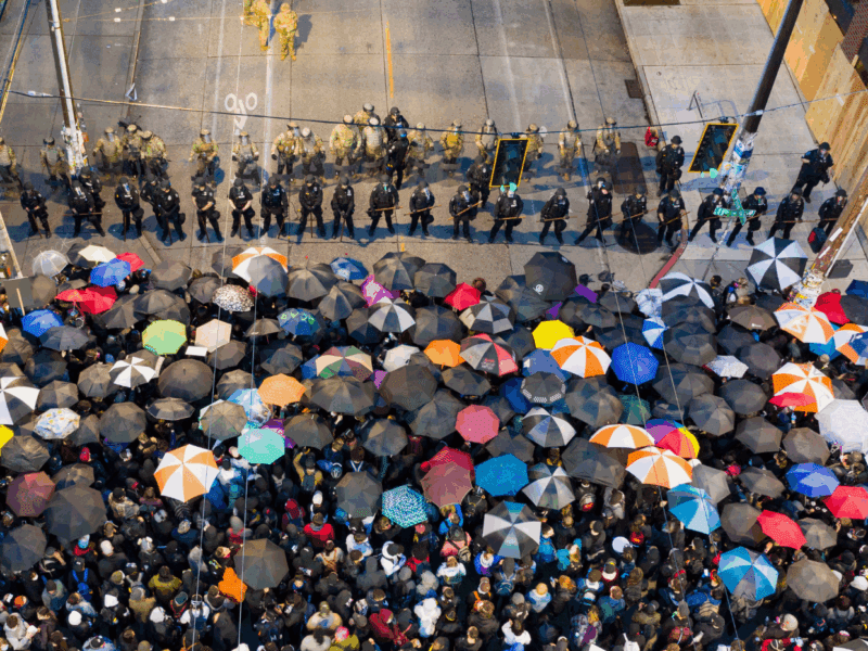 Protesters stand in the rain with umbrellas facing a line of armed riot police